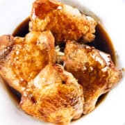 A white bowl holds four pieces of crispy skin chicken thighs, glazed to perfection. Coated in a rich bourbon glaze, the glossy sheen enhances their golden-brown allure. The light marble surface in the background complements this delicious chicken recipe.