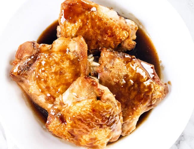 A white bowl holds four pieces of crispy skin chicken thighs, glazed to perfection. Coated in a rich bourbon glaze, the glossy sheen enhances their golden-brown allure. The light marble surface in the background complements this delicious chicken recipe.