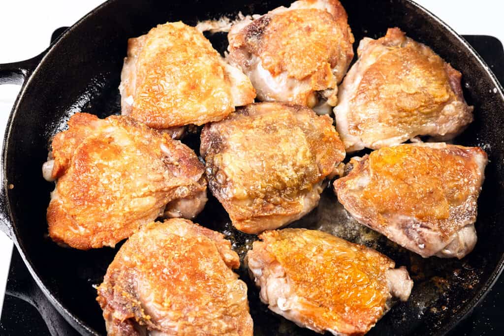 Close-up of a cast iron skillet containing eight golden-brown, crispy chicken thighs with a hint of bourbon glaze. The skin is perfectly seared and textured, suggesting they are being pan-fried or roasted. The surface sizzles slightly, highlighting their hot and freshly cooked state.