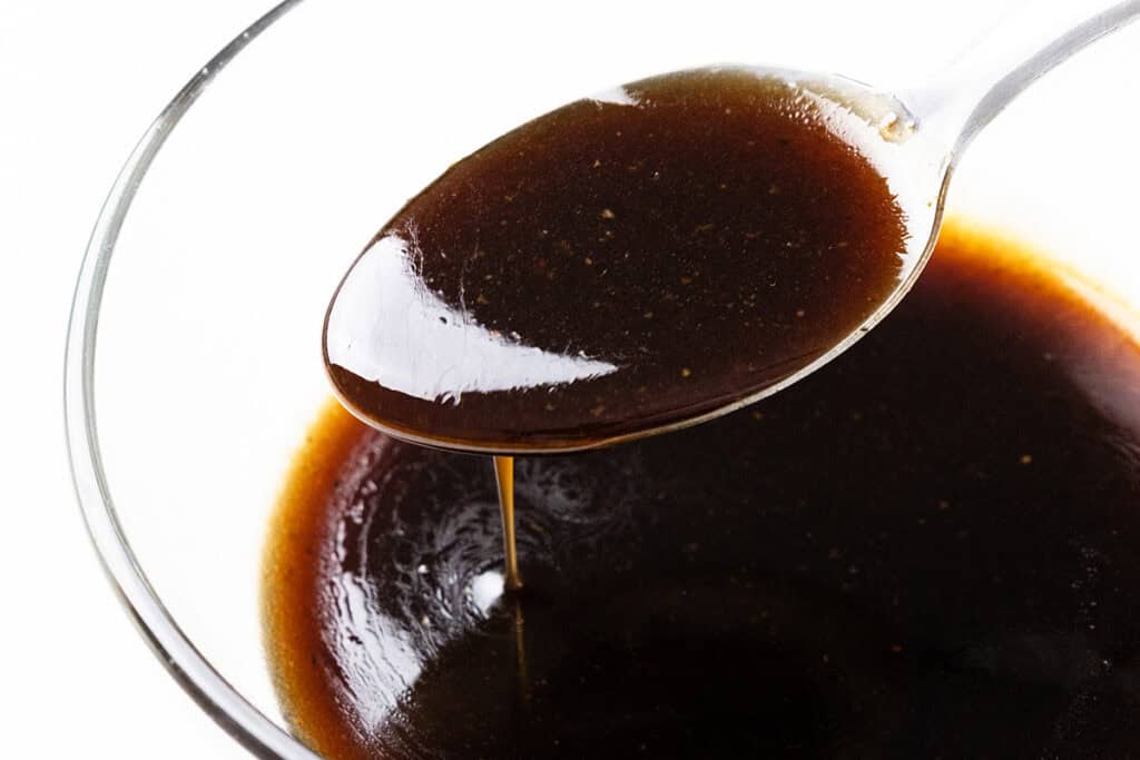 A close-up of a spoon holding a thick, dark brown bourbon glaze over a bowl filled with the same sauce. The glossy, smooth glaze boasts a rich consistency, perfect for drizzling over savory dishes like crispy skin chicken thighs.