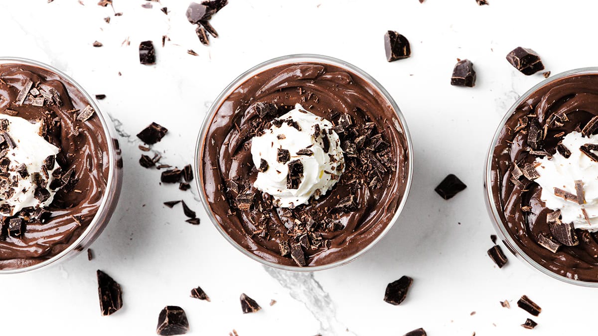 Top view of three glass cups filled with luscious chocolate pudding, each crowned with whipped cream and delicate chocolate shavings. Arranged on a white marble surface sprinkled with chocolate pieces, this delightful scene is perfect for any chocolate recipe collection.