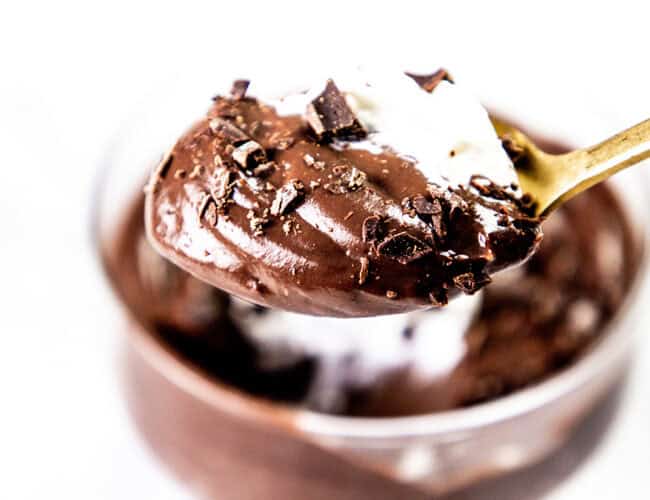 A close-up of a spoonful of creamy chocolate mousse topped with delicate chocolate shavings, showcasing the elegance of fine chocolate recipes. The mousse is smooth and rich, bits of chocolate atop it, set against a blurred backdrop of a glass filled with more mousse.