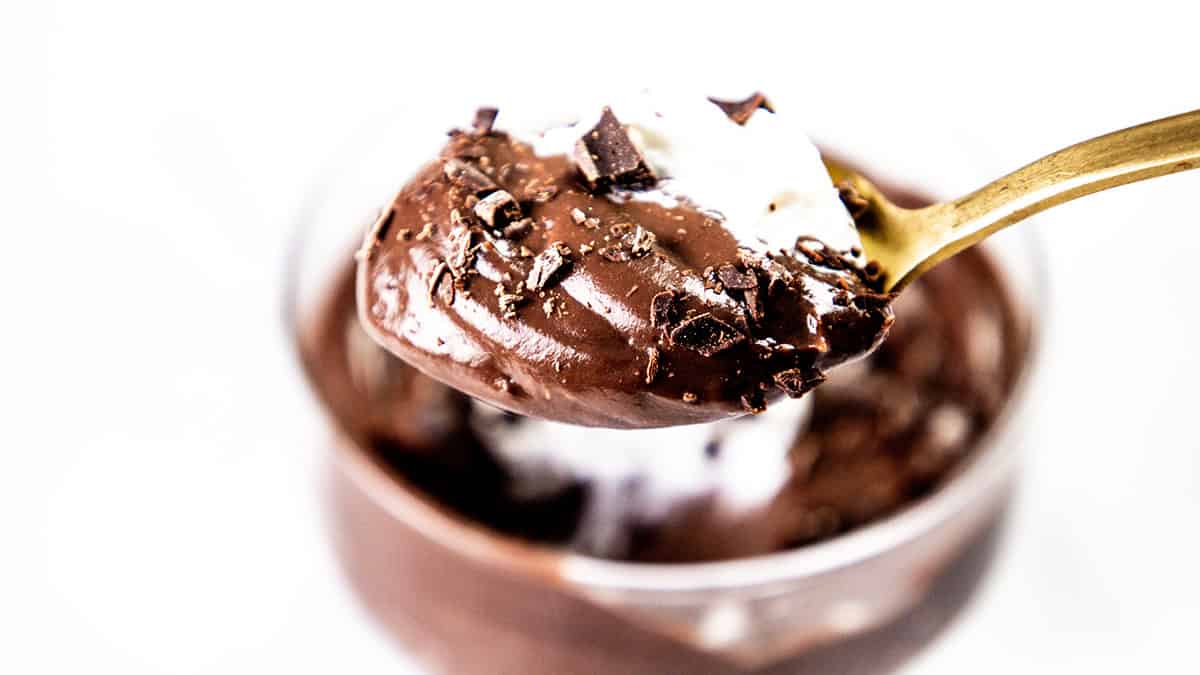 A close-up of a spoonful of creamy chocolate mousse topped with delicate chocolate shavings, showcasing the elegance of fine chocolate recipes. The mousse is smooth and rich, bits of chocolate atop it, set against a blurred backdrop of a glass filled with more mousse.