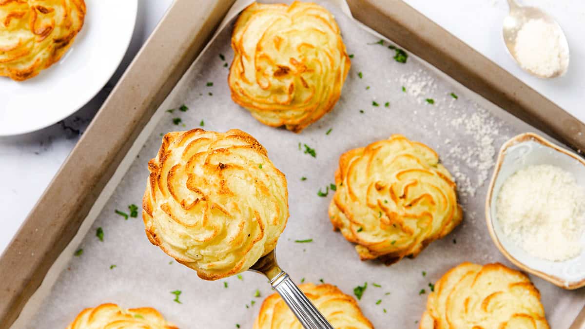 A baking tray lined with parchment paper holds several golden-brown duchess potatoes, a classic among sheet pan recipes. One is held up on a spatula, showcasing its perfectly crisp swirl. A small bowl of grated cheese and sprinkles of fresh herbs are artfully arranged around the tray.