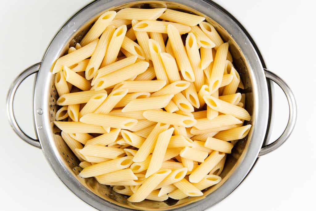 A stainless steel colander filled with cooked penne pasta, set against a white background. The pale yellow pasta sits alongside slices of savory olives, with some noodles standing upright, creating a delightful medley to complement any ricotta pesto dish.