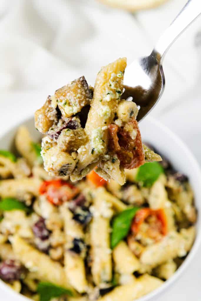 A close-up of a fork holding pasta with zucchini, tomatoes, olives, and cheese. This Eggplant Pasta dish is garnished with basil and appears creamy and seasoned with Ricotta Pesto, served in a white bowl in the background.