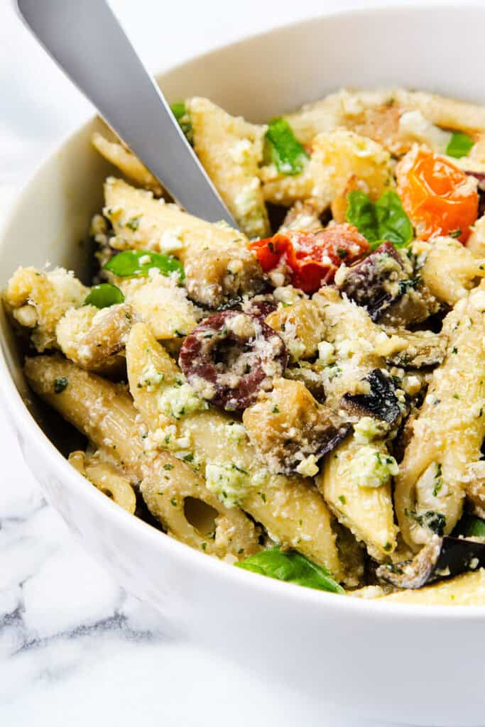 A bowl of creamy pasta with penne, vibrant cherry tomatoes, spinach, and mushroom slices, topped with grated cheese and a hint of ricotta pesto. A fork is digging into the pasta on the white marble surface, indicating a ready-to-eat meal.