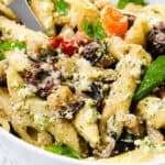 A close-up of a bowl of creamy pasta salad featuring penne, spinach, cherry tomatoes, olives, and mushrooms with a hint of ricotta pesto. The dish is vibrant and colorful, with a sprinkle of cheese and a knife partially visible in the bowl.