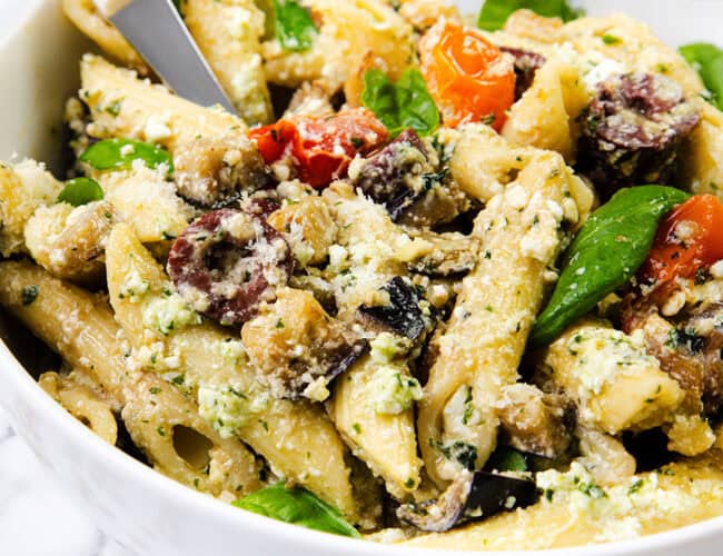 A close-up of a bowl of creamy pasta salad featuring penne, spinach, cherry tomatoes, olives, and mushrooms with a hint of ricotta pesto. The dish is vibrant and colorful, with a sprinkle of cheese and a knife partially visible in the bowl.