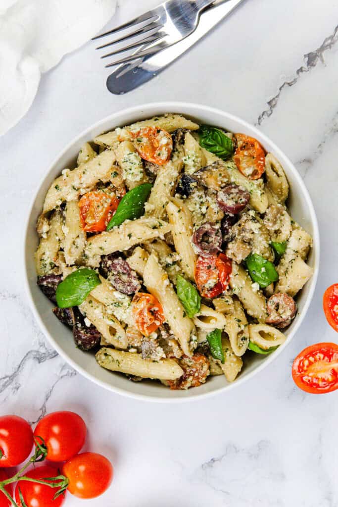 A bowl of eggplant pasta tossed with ricotta pesto, cherry tomatoes, black olives, and fresh basil leaves. A fork and knife rest on a white napkin nearby. A few cherry tomatoes are scattered at the bottom left corner on a marble surface.