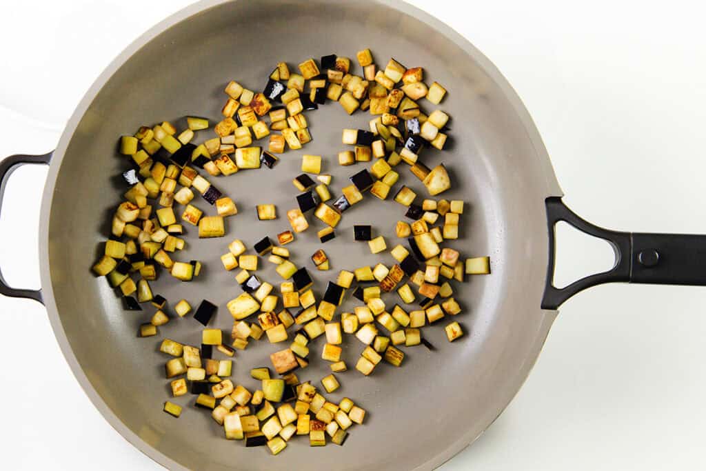 A frying pan with evenly sautéed small cubes of eggplant, golden brown and perfectly cooked. Scattered olives enhance the scene, their rich hues contrasting the white background. The pan's black handle adds a sleek touch to this culinary display.