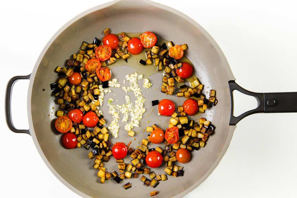 A frying pan containing diced eggplant, halved cherry tomatoes, and minced garlic, all sautéed together. Olives add a briny touch as the ingredients are evenly distributed around the pan.