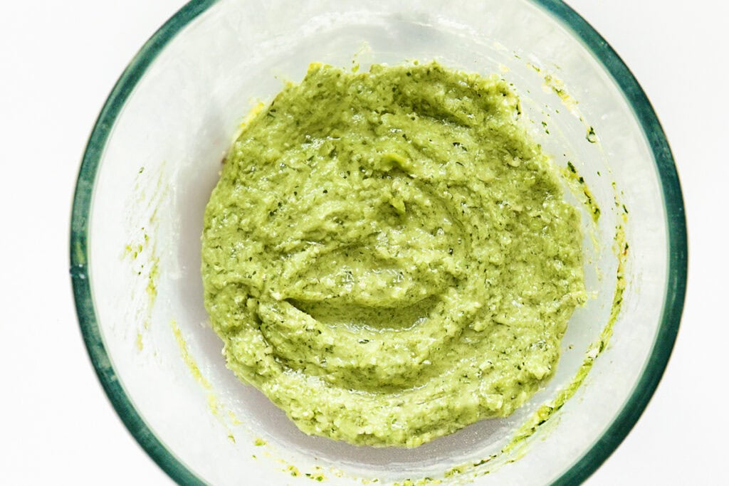 A glass bowl containing a creamy, green ricotta pesto sauce with visible herb flecks. The sauce has a smooth, slightly grainy texture. Perfect for drizzling over eggplant pasta or garnishing with olives, the bowl is placed against a plain white background.