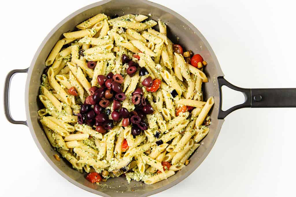 A skillet filled with penne pasta mixed with ricotta pesto, cherry tomatoes, and crumbled cheese, topped with a pile of black olives. The skillet has a handle on the right side.