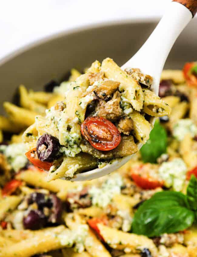 A serving spoon lifts a portion of Ricotta Pesto pasta with cherry tomatoes, olives, and pieces of eggplant from a larger bowl. The dish is garnished with fresh basil leaves.