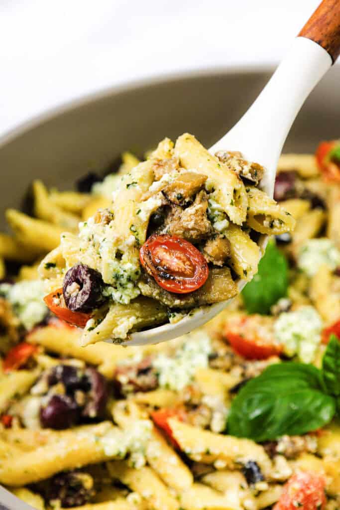 A serving spoon lifts a portion of Ricotta Pesto pasta with cherry tomatoes, olives, and pieces of eggplant from a larger bowl. The dish is garnished with fresh basil leaves.