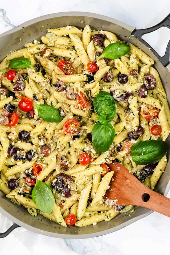 A skillet filled with penne pasta tossed in ricotta pesto, cherry tomatoes, black olives, and garnished with fresh basil leaves. A wooden spatula rests in the skillet, ready for serving.