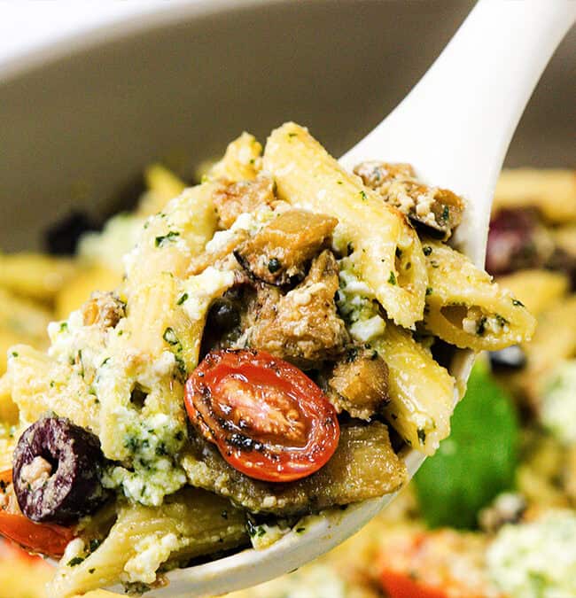 A spoonful of pasta with penne, cherry tomatoes, olives, mushrooms, and herbs, topped with ricotta cheese captures the essence of Mediterranean recipes. In the background, a bowl brimming with more of this delightful pasta dish awaits.