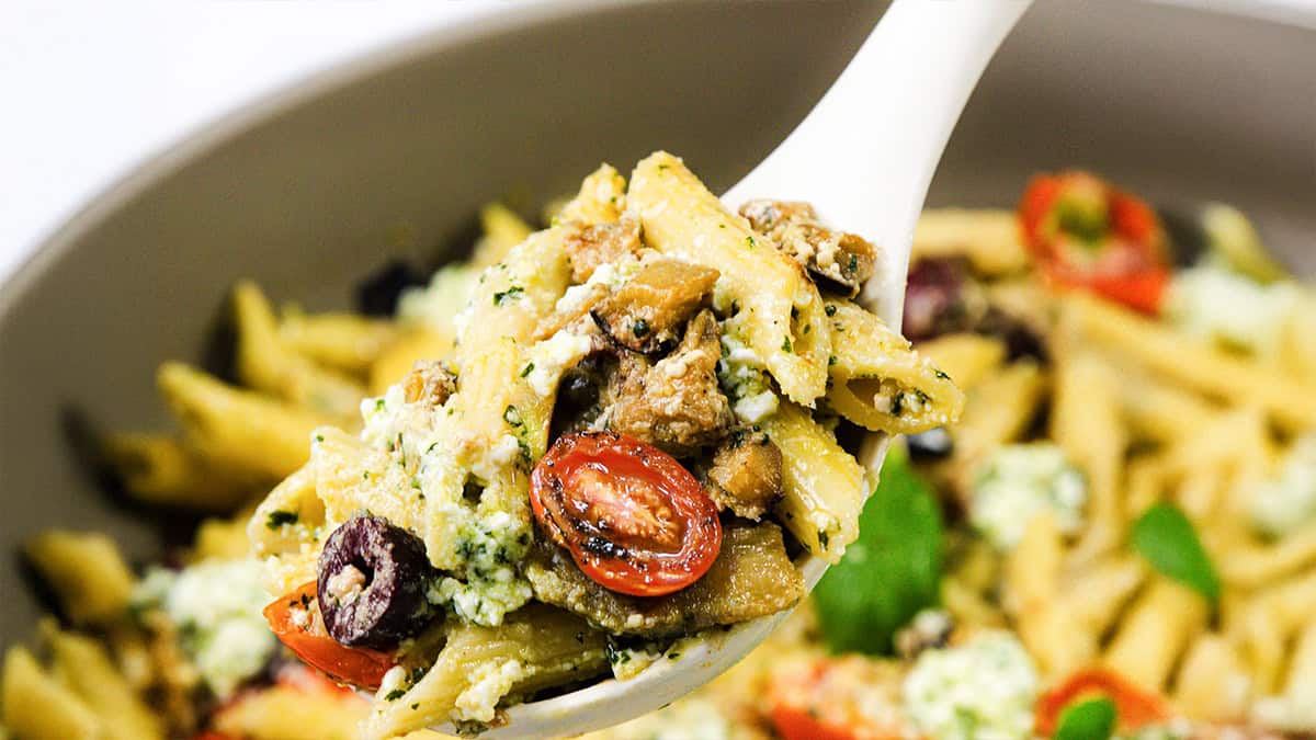 A spoonful of pasta with penne, cherry tomatoes, olives, mushrooms, and herbs, topped with ricotta cheese captures the essence of Mediterranean recipes. In the background, a bowl brimming with more of this delightful pasta dish awaits.