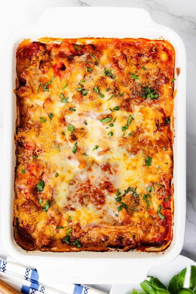 A baked lasagna casserole in a white rectangular dish, topped with melted cheese and sprinkled with fresh basil. The lasagna layers feature rich tomato sauce with hints of Italian sausage peeking through. A portion of a blue and white checkered cloth is visible at the bottom.