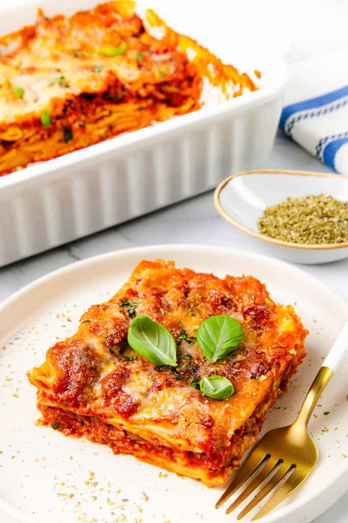 A slice of lasagna with layers of pasta, Italian sausage, tomato sauce, and melted cheese is garnished with fresh basil on a white plate. A fork rests beside it. In the background is a casserole dish filled with more lasagna and a small bowl of dried herbs.