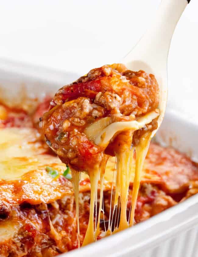 A serving of Italian Sausage Ravioli Casserole is being lifted from a white baking dish with a spoon. This hearty casserole is rich with melted cheese, layers of pasta, tomato sauce, and Italian sausage, with strings of cheese stretching from the dish.