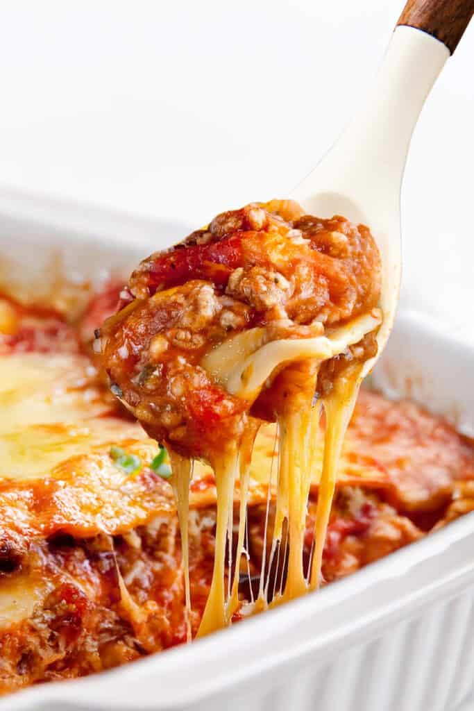 A serving of Italian Sausage Ravioli Casserole is being lifted from a white baking dish with a spoon. This hearty casserole is rich with melted cheese, layers of pasta, tomato sauce, and Italian sausage, with strings of cheese stretching from the dish.