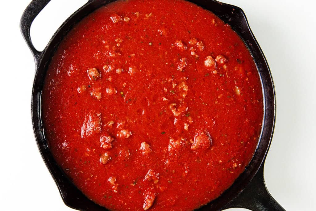 A cast iron skillet brimming with rich, chunky red tomato sauce and Italian sausage, featuring visible pieces of tomato and herbs, on a white background.