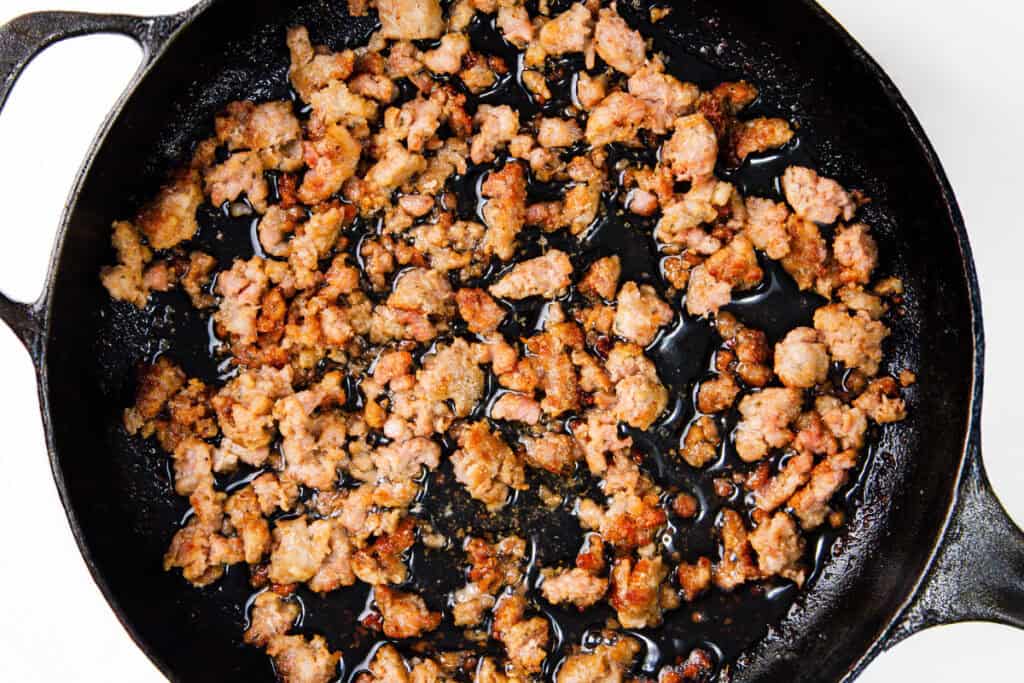 Crumbled and browned Italian sausage cooks in a black cast iron skillet, the meat glistening from the heat and oils. The skillet sits on a light surface, highlighting the contrast between the rich, golden-brown sausage and the dark pan.