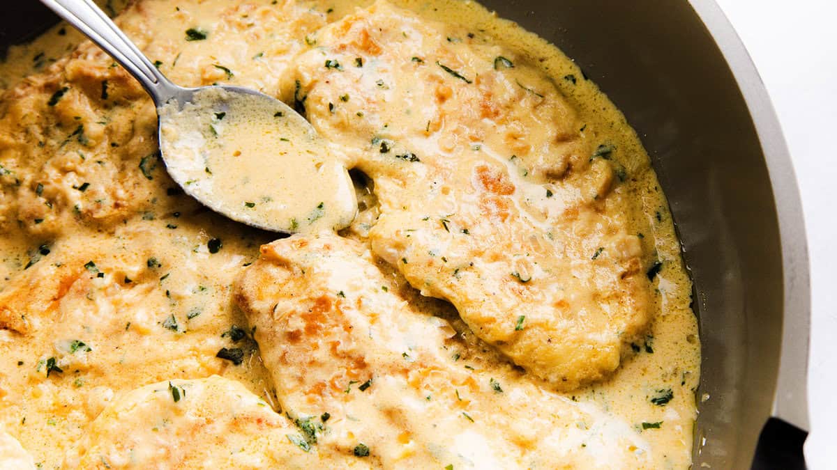 Creamy chicken breasts with a golden crust simmer in a rich, herb-infused sauce, offering restaurant-quality appeal. A silver spoon rests on the chicken in a pan, showcasing the dish's appetizing texture and color—a perfect meal to make at home.