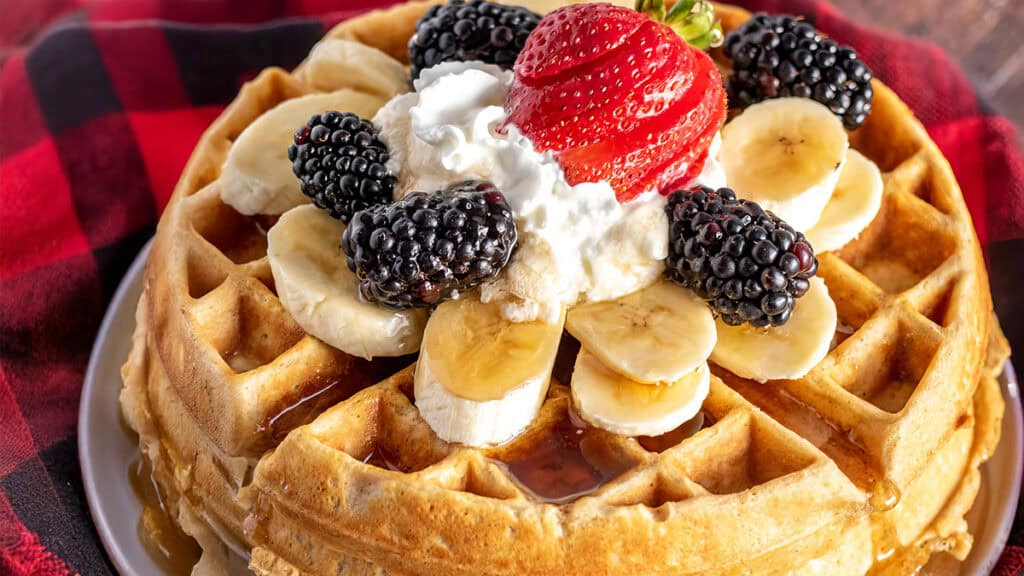 A stack of waffles, inspired by oat recipes, topped with sliced bananas, blackberries, a dollop of whipped cream, and a strawberry sits on a red and black checkered cloth. Syrup cascades down the sides in sweet perfection.