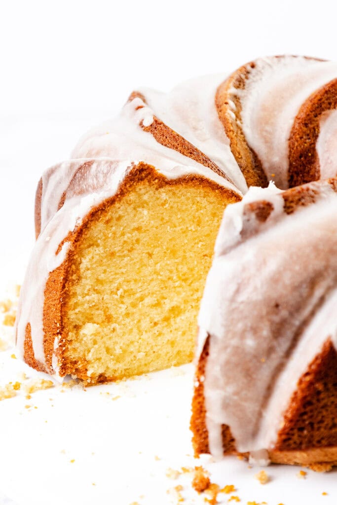 A close-up of a sliced citrus and olive oil bundt cake with a golden-brown crust and a light, moist interior. The cake is topped with a smooth white glaze, and a few crumbs are visible on the white surface beneath.