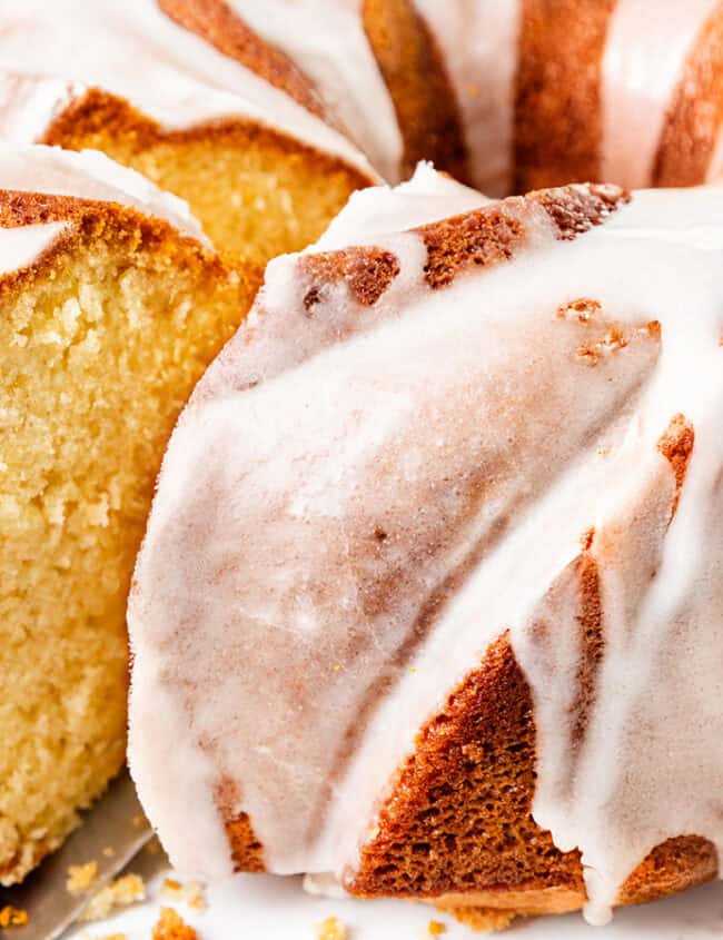 A close-up of a sliced bundt cake covered in white glaze. The cake appears moist and golden, with a smooth, shiny icing drizzled evenly over its textured surface.