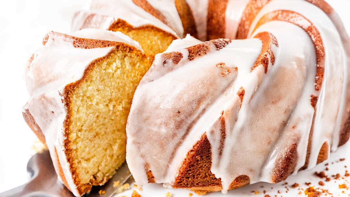 A close-up of a sliced bundt cake covered in white glaze. The cake appears moist and golden, with a smooth, shiny icing drizzled evenly over its textured surface.