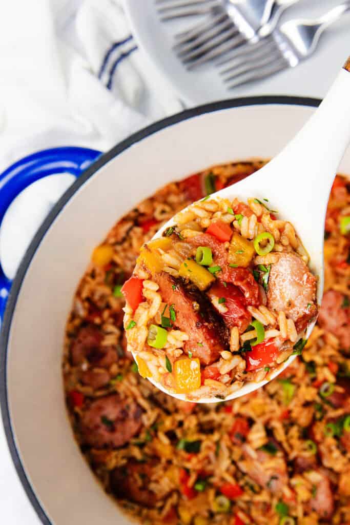 A spoonful of One Pot Sausage and Rice with sliced sausage, rice, diced tomatoes, and bell peppers is held above a blue pot filled with the flavorful one-pot dish. Green onions garnish the vibrant mixture, and a striped towel and silverware are visible in the background.