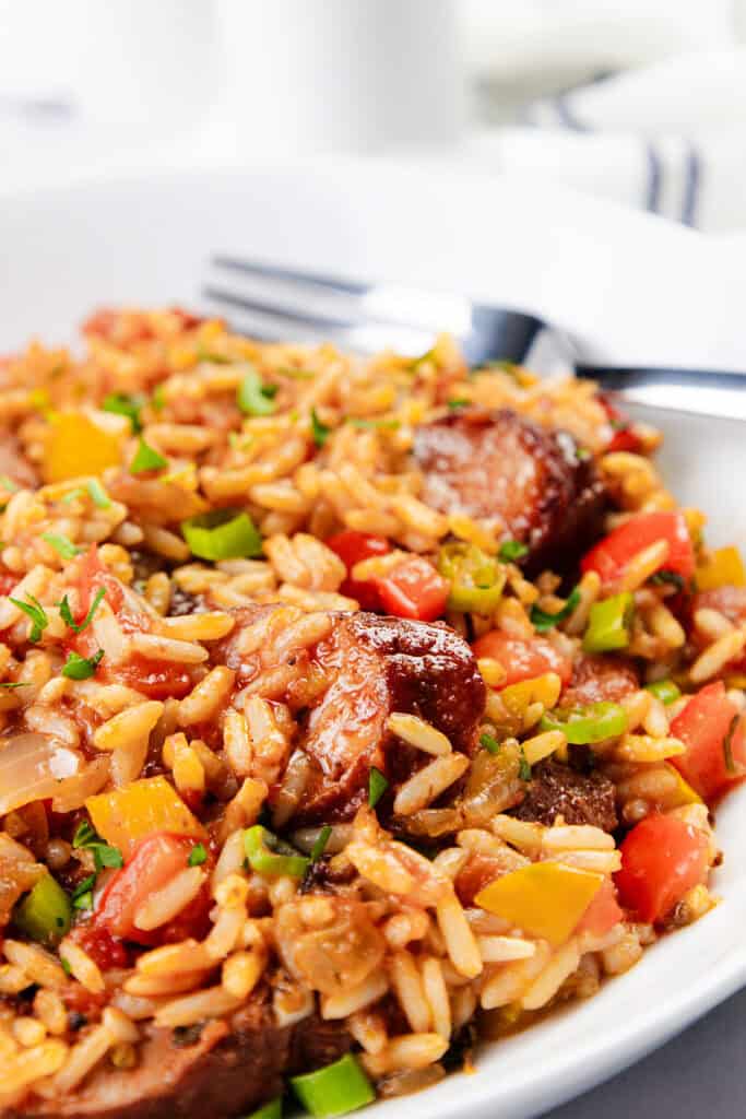 A close-up of a one-pot Jambalaya featuring rice, sliced sausage, bell peppers, and chopped green onions. The dish is richly colored with vibrant reds, yellows, and greens, beautifully presented on a white plate with a fork.