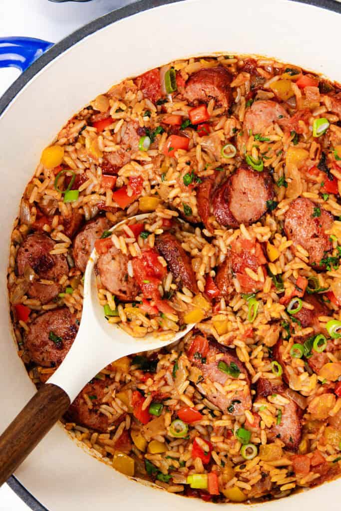 A One Pot Sausage and Rice brimming with sliced sausage, rice, and diced bell peppers in a rich tomato-based sauce. A white spoon scoops the savory mixture while green onions are sprinkled on top for garnish.