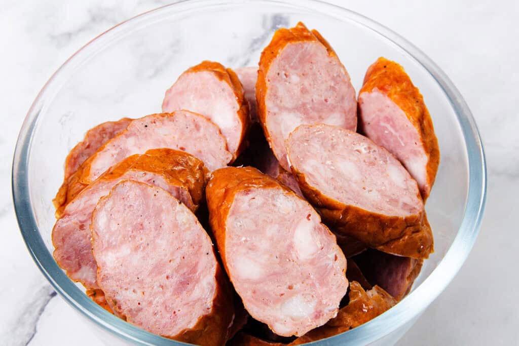 Slices of sausage are piled in a clear glass bowl, placed on a white marble surface. The sausages have a pinkish interior with a reddish-brown casing, creating the perfect start to an irresistible one-pot dish.