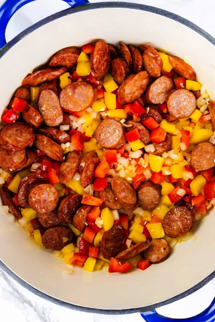 A pot containing sliced sausages, diced red and yellow bell peppers, and chopped onions cooking.