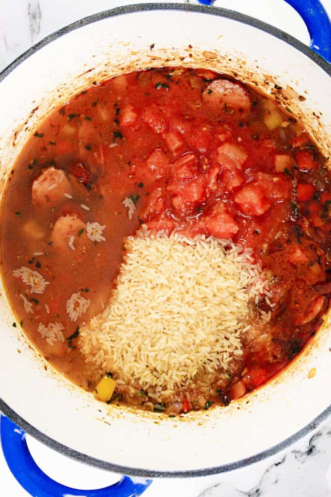 A one-pot meal featuring a tomato-based stew brimming with sausage chunks and tender pieces of meat. A mound of uncooked rice crowns the dish, seasoned with aromatic herbs. The pot, accented by blue handles, rests elegantly on a marble surface.