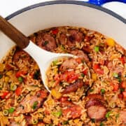 A pot of One Pot Sausage and Rice filled with flavorful featuring sliced sausages, colorful bell peppers, and rice. A white spoon lifts a portion, showcasing the vibrant mix of ingredients. The pot has a blue rim and a wooden handle.