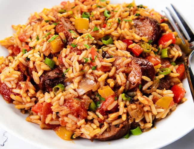 A plate of sausage and rice featuring Southern Recipes flair, with rice, sausage, peppers, tomatoes, and green onions garnished with herbs. A fork is placed to the side on a white surface.