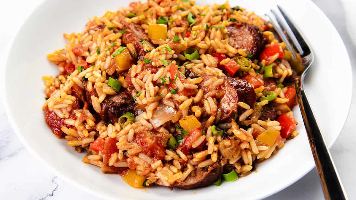 A plate of sausage and rice featuring Southern Recipes flair, with rice, sausage, peppers, tomatoes, and green onions garnished with herbs. A fork is placed to the side on a white surface.