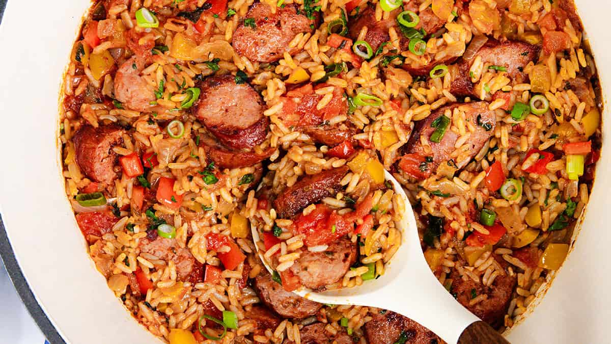 A vibrant one pot recipe, this sausage and rice is a feast for the senses, boasting sliced sausage, rice, diced tomatoes, bell peppers, and chopped green onions. The dish is presented in a white pot with a ladle ready to serve up deliciousness.