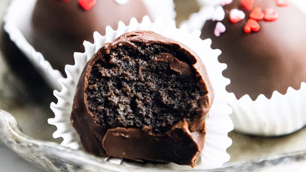 Close-up of Oreo truffles nestled in white paper cups. One truffle in the foreground has a bite taken out, revealing a rich, dark interior. Decorated with red and pink heart sprinkles, these easy dessert recipes add a delightful twist to classic treats.