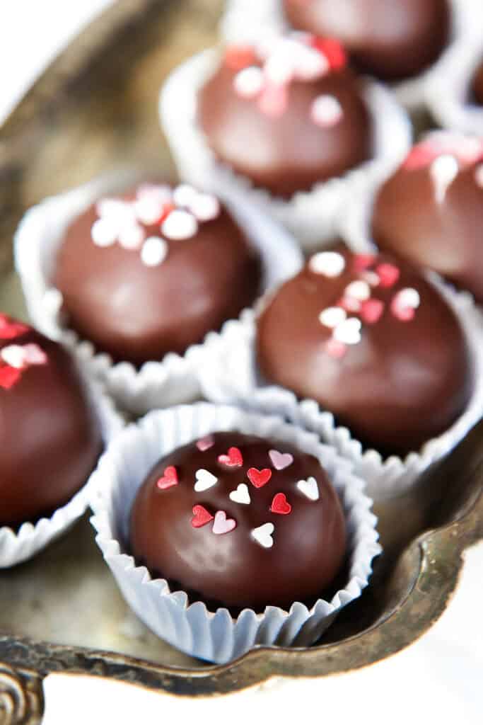 A tray of Oreo truffles in paper liners, decorated with red and white heart-shaped sprinkles. The truffles are arranged neatly, showcasing a glossy, smooth surface.