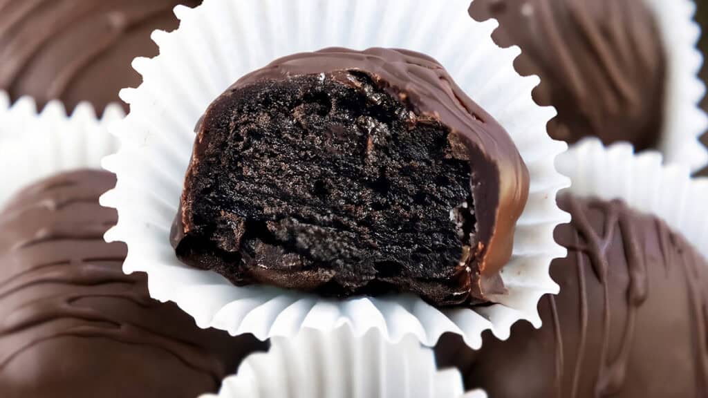 Close-up of a chocolate truffle, bitten to reveal a dark, rich filling—a creation straight from gourmet chocolate recipes. It's placed in a white paper cup with other similar truffles in the background. The surface of the chocolate is smooth and glossy, with a drizzle on top.