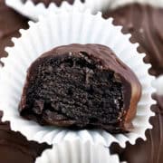 A close-up of an Oreo truffle with a bite taken out reveals a dark, rich interior speckled with cookie crumbs. It sits in a crinkled white paper wrapper, while other decadent dessert truffles are blurred in the background.