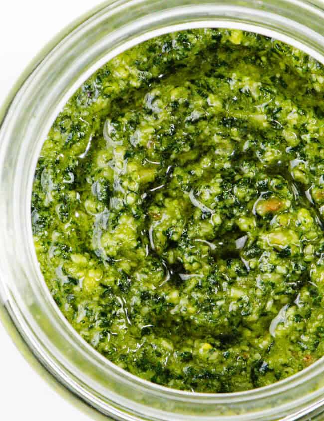 A close-up of a jar filled with homemade pesto sauce. The pesto has a textured, oily surface with visible herbs and nuts, giving it a fresh and vibrant appearance. This quick and easy recipe highlights its rich green color and slightly chunky consistency.