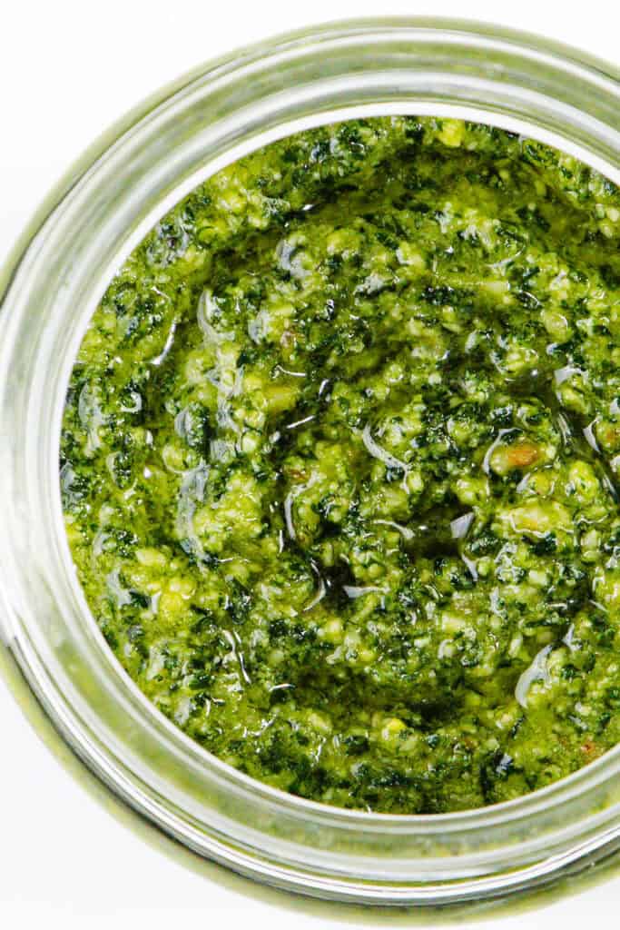 A close-up of a jar filled with homemade pesto sauce. The pesto has a textured, oily surface with visible herbs and nuts, giving it a fresh and vibrant appearance. This quick and easy recipe highlights its rich green color and slightly chunky consistency.